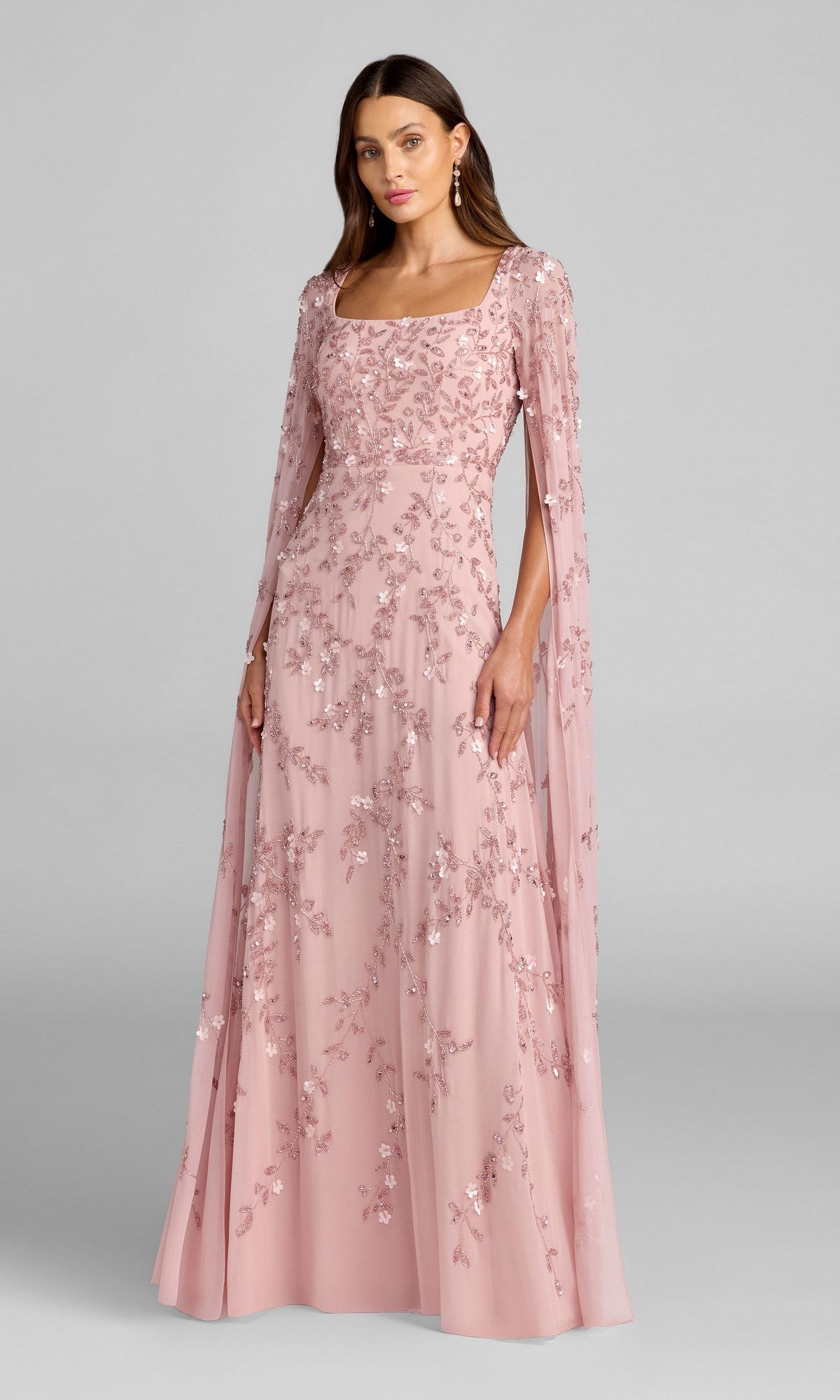 A woman wearing the Mac Duggal 23068 long formal dress in rose, featuring floral embroidery and cape sleeves, poses against a plain gray background.